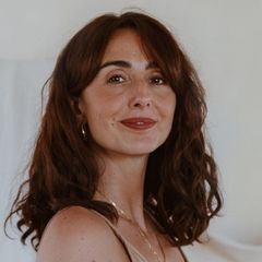The image features a woman with wavy, medium-length brown hair. She is wearing a light-colored, sleeveless top and has a subtle smile on her face. Her jewelry includes a bracelet and small hoop earrings. The background is softly blurred with neutral tones, possibly featuring draped fabric.