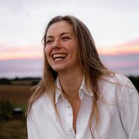 The image features a person with long hair, smiling and laughing. They are wearing a white button-up shirt. The background shows a landscape with fields and a colorful sky, indicating either sunrise or sunset, with shades of pink and purple. Overall, the scene conveys a cheerful and relaxed atmosphere.