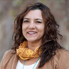 The image features a woman with curly hair, smiling at the camera. She is wearing a light-colored top with a brown jacket and a yellow scarf wrapped around her neck. The background is blurred with soft colors, suggesting an outdoor setting.