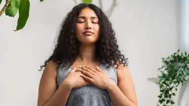 The image shows a woman with curly hair, standing with her eyes closed and a calm expression. She has her hands placed over her heart and appears to be in a moment of reflection or meditation. The background features greenery, suggesting a peaceful indoor environment.