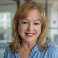 The image shows a woman with medium-length blonde hair and a light blue shirt. She is smiling and appears to be in a well-lit indoor setting, possibly an office. The background is blurred, but there are indications of a modern, professional environment.