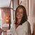 The image features a woman smiling while standing in a well-decorated interior space. She has long, dark hair with lighter highlights and is wearing a white shirt. The background includes elements of home décor, such as warm lighting, plants, and curtains, creating a cozy atmosphere. There are also decorative elements on the wall. The overall vibe of the image is welcoming and stylish.