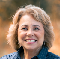The image features a woman with light-colored, wavy hair and a warm smile. She is wearing a blue shirt and appears to be outdoors, with a blurred background that suggests a natural setting, possibly with soft, warm colors. The expression on her face conveys happiness and friendliness.