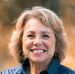 The image features a woman with light-colored, wavy hair and a warm smile. She is wearing a blue shirt and appears to be outdoors, with a blurred background that suggests a natural setting, possibly with soft, warm colors. The expression on her face conveys happiness and friendliness.
