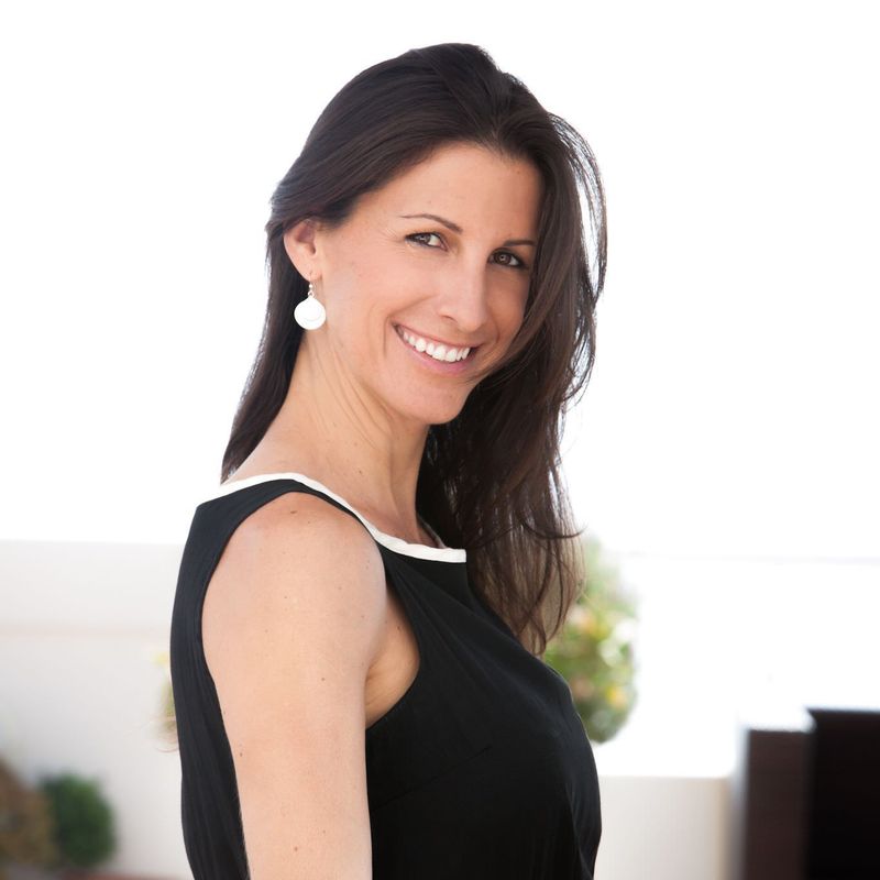The image shows a woman with long, dark hair, smiling and looking to the side. She is wearing a black dress with a contrasting white trim. The background appears bright, likely outdoors, with a hint of greenery and a blurred area that may indicate additional architectural elements.
