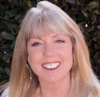 The image features a woman with long, straight blonde hair, smiling brightly. She is wearing a white sweater with a silver trim and a pendant necklace that has a blue-colored stone. The background consists of green foliage, which provides a natural setting.