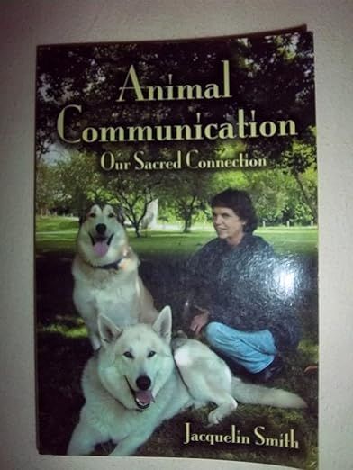 The image shows a book titled "Animal Communication: Our Sacred Connection." The cover features a person sitting on the ground with two large dogs in a grassy, park-like setting. The dogs appear to be content and are oriented towards the person. The background includes trees and a natural landscape. The author's name, Jacquelin Smith, is also visible on the cover.