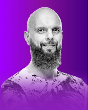 The image features a bald man with a beard, smiling at the camera. His attire has a colorful, artistic design. The background is a gradient of purple hues, which enhances the overall look of the image.