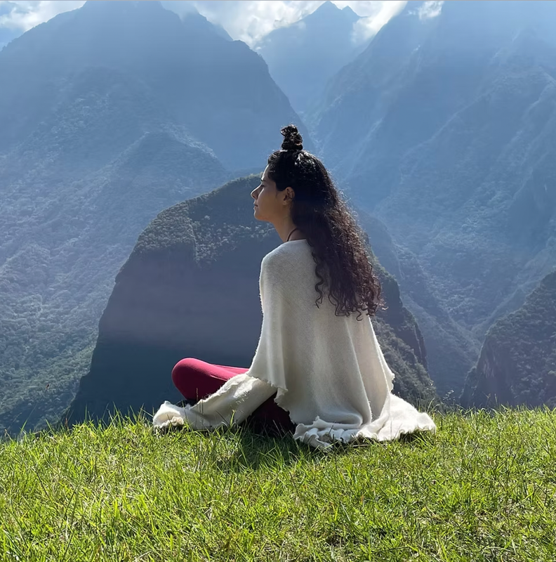 The image shows a person sitting on a grassy area, facing a scenic view of mountains. The individual is wearing a light sweater and red pants, with long, curly hair. The backdrop features steep, lush mountains and a blue sky with some clouds. The overall setting appears serene and natural, suggesting a tranquil outdoor environment.
