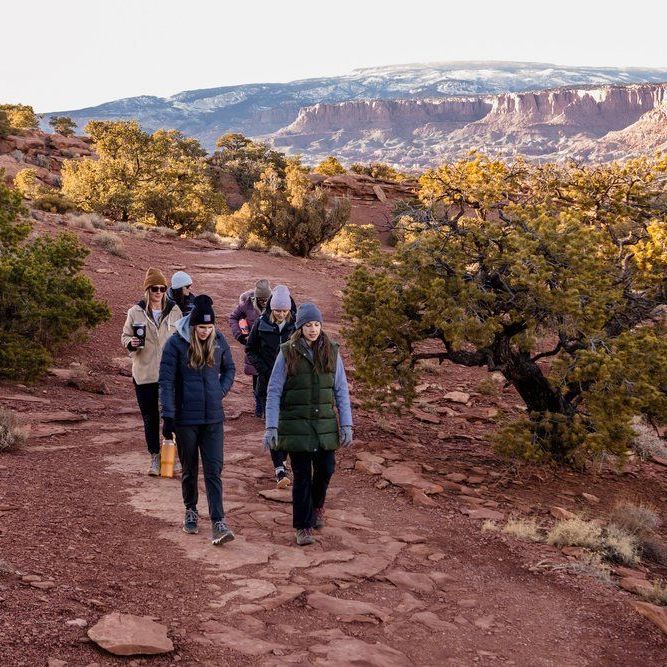 The image depicts a group of five people walking along a path in a rocky, outdoor landscape. The scenery features reddish-brown terrain, with sparse vegetation, including some trees. In the background, there are layered rock formations and hills. The group is dressed for cooler weather, wearing jackets and hats, and they appear to be engaged in outdoor activities, likely hiking. The light suggests it's early morning or late afternoon, casting warm tones across the landscape.