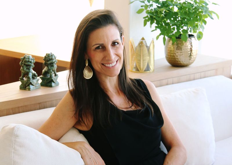 The image features a woman sitting on a white couch. She has long, dark hair and is wearing a black dress. The setting appears to be a well-lit interior space with a light background. There are decorative elements nearby, including green plants in pots and small statues on a table in the background. The atmosphere looks relaxed and inviting.