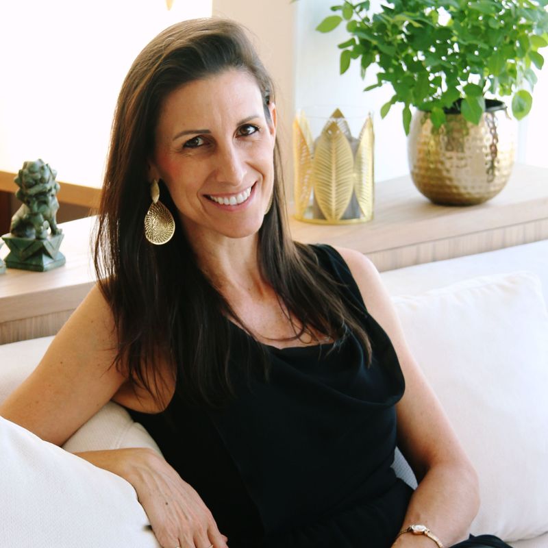 The image features a woman sitting on a white couch. She has long, dark hair and is wearing a black dress. The setting appears to be a well-lit interior space with a light background. There are decorative elements nearby, including green plants in pots and small statues on a table in the background. The atmosphere looks relaxed and inviting.