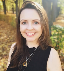 The image features a person smiling in an outdoor setting. The background is filled with autumn foliage, with leaves scattered on the ground. The individual has long, brown hair and is wearing a sleeveless, black top. They also have a necklace with a pendant. The lighting appears warm and natural, suggesting a sunny day.