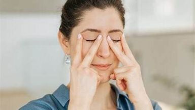 The image shows a woman with her eyes closed, using her fingers to gently apply pressure around her eyes. She is wearing a blue shirt and appears to be in a calm, relaxed indoor setting. The atmosphere looks bright and cozy. The woman seems focused, likely engaging in a relaxation technique or facial exercise.