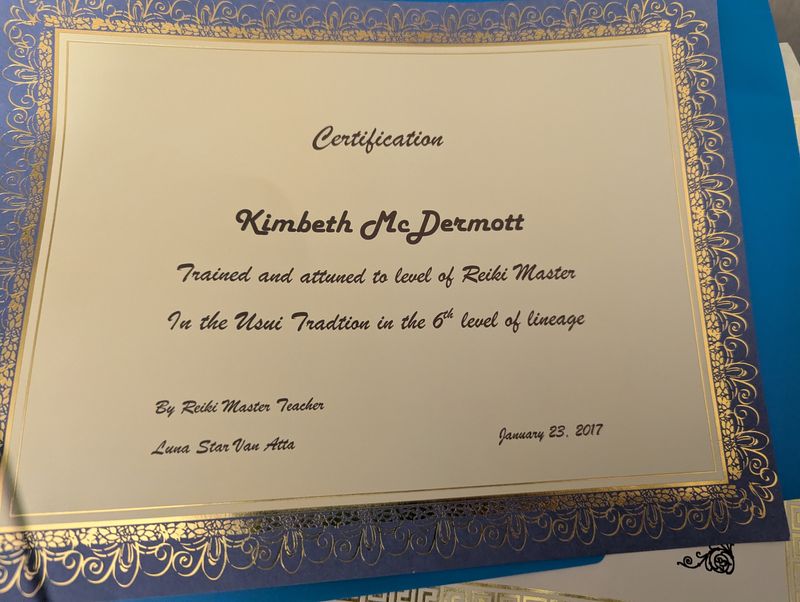 The image shows a certification document. It is decorated with gold embellishments and has a formal layout. The text indicates that the individual named is trained and attuned to the level of Reiki Master, specifically in the Usui Tradition at the 6th level of lineage. The certification is signed by a Reiki Master Teacher and is dated January 23, 2017. The background is a blue surface, and the document has ornate borders.