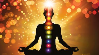 The image depicts a silhouette of a person sitting in a meditative pose, with their legs crossed and hands resting on their knees. The figure is outlined against a vibrant, warm-colored background filled with soft, glowing orbs of light. Along the center of the silhouette, there are seven colored circles representing the chakras, from the base of the spine to the crown of the head, with a bright light or glow emanating from the top of the head. The overall theme conveys a sense of tranquility, spirituality, and energy.