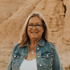 The image shows a smiling woman standing outdoors. She has long, light brown hair and is wearing glasses. She is dressed in a denim jacket over a white top. The background features a natural landscape with rocky formations, suggesting a desert or arid environment.