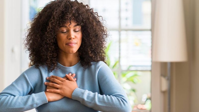 The image shows a woman sitting at a table with her eyes closed and hands placed over her heart. She has curly hair and is wearing a light blue sweater. The setting appears to be indoors, with soft lighting and a background that includes a window and some greenery visible outside. The overall mood of the image suggests a moment of calmness or meditation.