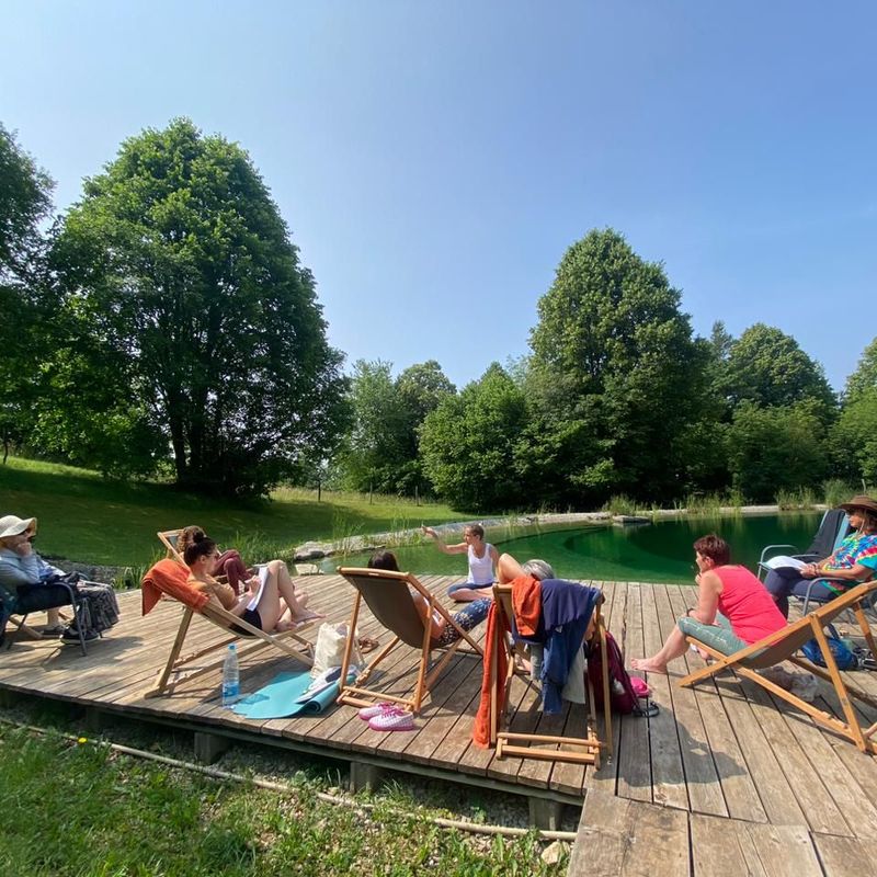 The image shows a group of people sitting on a wooden deck by a body of water, likely a pond or lake, surrounded by greenery. They are lounging in deck chairs and appear to be engaged in conversation or relaxation. The setting is bright and sunny, with trees in the background and a clear blue sky. Some of the individuals are wearing hats, and there are personal items like a water bottle and clothing scattered around.
