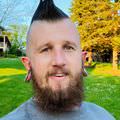 The image shows a man with a distinctive hairstyle featuring a mohawk. He has a full beard and large ear gauges. He is smiling and standing outdoors in a green area with trees in the background. The lighting suggests it's a sunny day. The man is wearing a gray shirt.