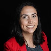 The image features a woman smiling against a black background. She has long, dark hair and is wearing a bright red blazer over a patterned top. The overall expression is friendly and approachable.
