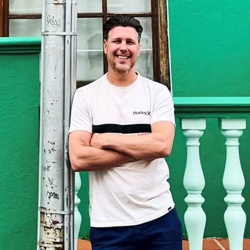 The image shows a man standing by a green building. He is leaning against a utility pole with his arms crossed. The building features a large window and a door, with a number "140" visible on the wall. There are white railings next to the man and a terracotta-tiled pathway leading up to the entrance. The man is wearing a light-colored t-shirt and dark shorts, paired with white shoes. The overall scene appears to be bright and casual.