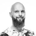 The image is a black-and-white portrait of a man with a bald head and a thick beard. He is smiling and has crossed arms. He is wearing a patterned shirt that features abstract designs in various colors. The background is plain and light, adding focus to his expression and attire.