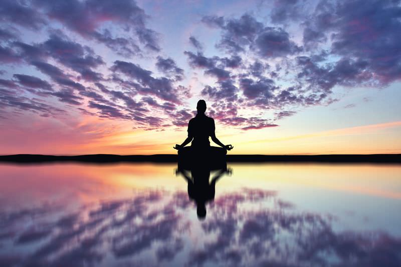 The image depicts a silhouette of a person meditating in a cross-legged position. This figure is seated in front of a calm body of water, which reflects both the figure and the vibrant colors of the sky. The sky is filled with a gradient of hues ranging from soft pink and orange to deep blue, accompanied by clouds. The overall atmosphere conveys a sense of tranquility and serenity.