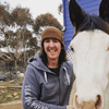 The image shows a person standing next to a white horse. The person is smiling and wearing a grey hoodie and a brown beanie. The background consists of a blue building and a staircase. There are some trees in the area, and the ground appears to be dirt. The horse is looking directly at the camera.