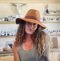 The image shows a person with long, curly hair wearing a wide-brimmed orange hat and a gray tank top. The background features shelves with various bottles and jars, suggesting a store or workshop setting. There appears to be a decorative element on the wall above the shelves, and the overall atmosphere seems warm and inviting.