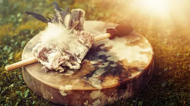 The image shows a round wooden drum resting on a grassy surface. On top of the drum, there is a bundle of herbs or sage wrapped in a cloth, adorned with feathers. A stick, likely used for smudging, rests alongside the bundle. Soft lighting enhances the natural setting.