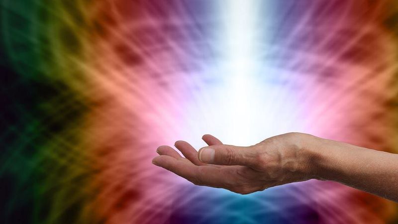 The image shows an open hand reaching out, with a vibrant and colorful background featuring swirling patterns in hues like purple, green, orange, and pink. In the center of the image, there is a bright, white light emanating, creating a radiant effect that suggests energy or illumination. The overall composition gives a sense of depth and movement.