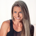 The image shows a person with long, straight blonde hair and bright blue eyes. They are smiling and wearing a black tank top. The background is light-colored and softly diffused, suggesting a well-lit environment.