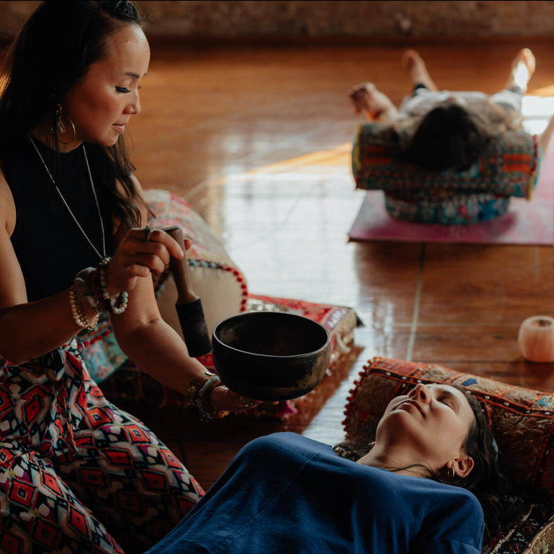 The image shows a wellness or healing session taking place in a serene setting. In the foreground, a woman is lying on a decorative cushion, eyes closed, appearing relaxed. Another woman is seated beside her, holding a singing bowl and seemingly preparing to make sound for therapeutic purposes. In the background, another person is lying on their back on a separate cushion. Various candles and small objects are visible nearby, contributing to the tranquil atmosphere. The lighting suggests a warm, inviting space, ideal for relaxation and healing activities.
