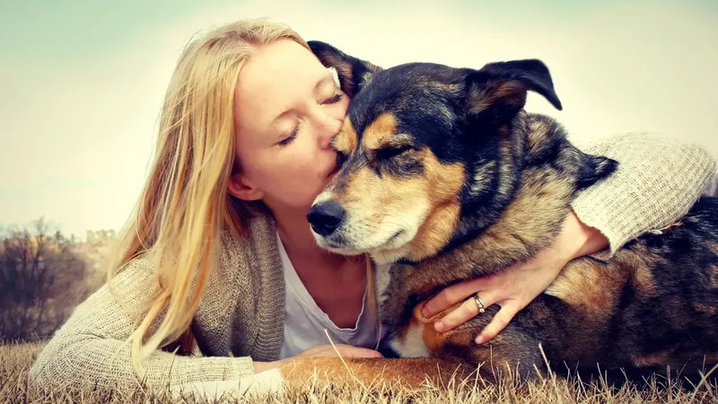 The image shows a person with long blonde hair, leaning down to kiss a dog. The dog appears to be a medium-sized, fluffy breed with a dark coat and lighter markings. They are lying on a grassy area, and the background features a blurred landscape. The scene conveys a sense of affection and companionship between the person and the dog.