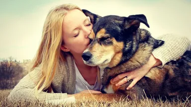 The image shows a person with long blonde hair, leaning down to kiss a dog. The dog appears to be a medium-sized, fluffy breed with a dark coat and lighter markings. They are lying on a grassy area, and the background features a blurred landscape. The scene conveys a sense of affection and companionship between the person and the dog.