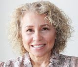 The image features a woman with curly hair, smiling. She appears to be wearing a patterned top and has a warm expression. The background is light and neutral.