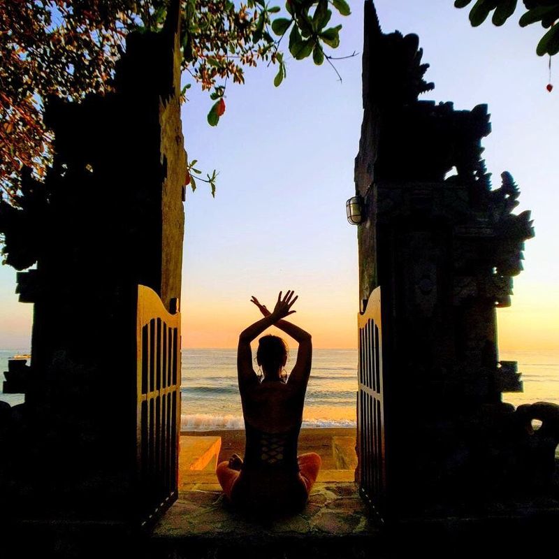 The image shows a silhouette of a person sitting on the ground, framed between two ornate stone structures that resemble gates or entranceways. The individual has their arms raised above their head, creating a peaceful pose. The background features a beach at sunset, with gentle waves lapping at the shore, and the sky displaying vibrant colors as the day transitions to night. There are also some greenery and leaves visible, adding to the serene atmosphere of the scene.