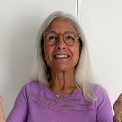 The image features a woman with gray hair, wearing a light purple long-sleeve shirt. She is smiling and gesturing with both hands, holding them out to her sides. The background is a plain white wall. Her glasses are stylish, and she appears to be in a cheerful and expressive mood.