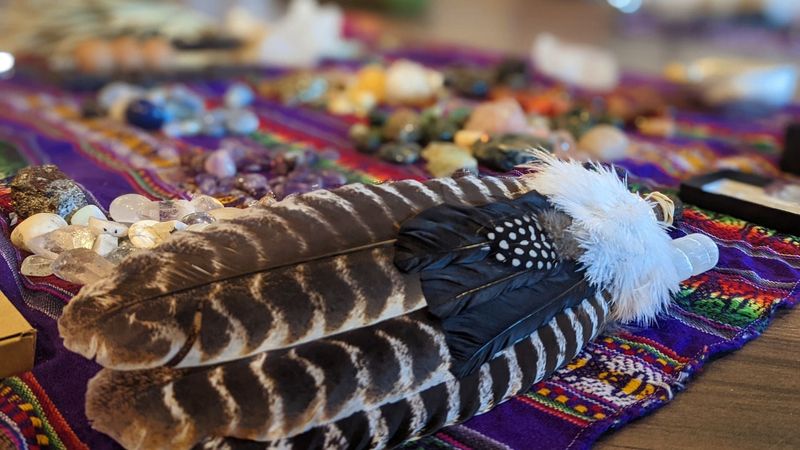 The image features a vibrant textile, likely a woven cloth, with a variety of colorful objects scattered across it. Prominently displayed in the foreground are two large feathers, decorated with a mix of browns and whites, and a smaller black feather with white spots. Surrounding these feathers are various gemstones and crystals, showcasing an array of colors, along with some additional objects that are difficult to identify. The overall setting seems to suggest a spiritual or ritualistic context.