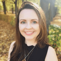 The image features a person with long brown hair, smiling at the camera. She is wearing a black sleeveless top and a necklace with a geometric pendant. The background shows a park or natural setting with trees and scattered autumn leaves on the ground, indicating a warm, sunny day.