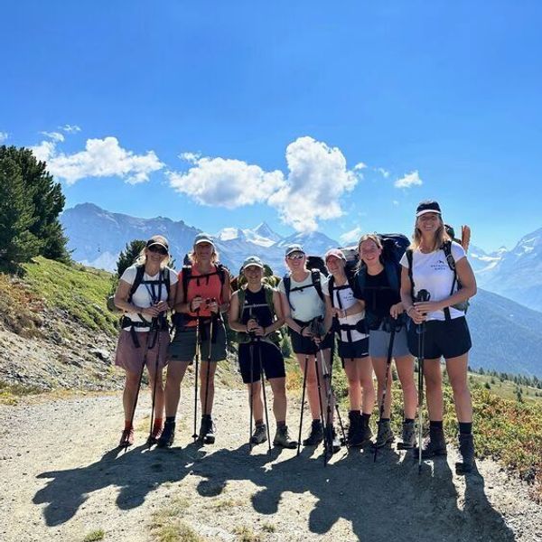 The image shows a group of seven individuals standing together on a hiking trail. They are wearing outdoor clothing suitable for hiking, including shorts and backpacks. Each person is holding hiking poles. In the background, there are mountainous landscapes and a clear blue sky with some clouds. The scenery looks lush and natural, indicating a beautiful day for hiking.