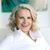 The image depicts a woman with short, wavy blonde hair, styled with an undercut on one side. She is wearing a white shirt and is smiling broadly. The background features abstract art with soft colors. The overall vibe of the image is cheerful and professional.