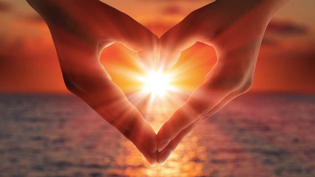 The image features two hands positioned to form a heart shape. In the center of this heart, there is a bright glow resembling the sun. The background shows a sunset over a serene body of water, with warm colors like orange and yellow reflecting on the surface. The overall composition conveys a romantic or peaceful atmosphere.