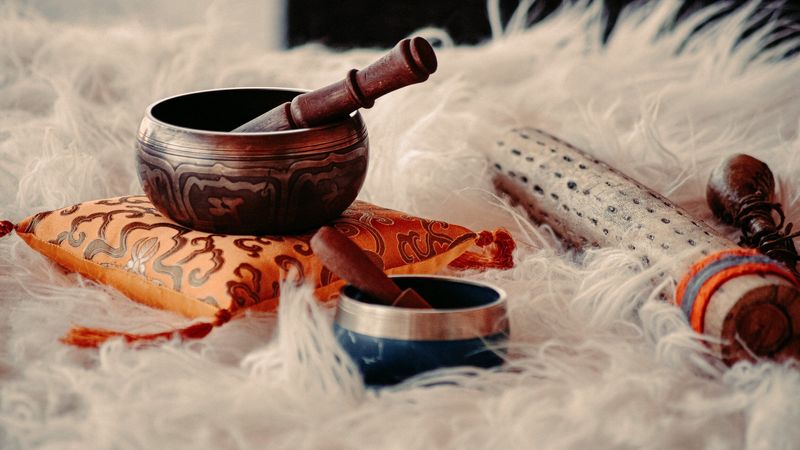 The image features a soft, fluffy white surface with a couple of small bowls. One bowl is dark and ornate, resting on an orange and patterned cushion, along with a mallet used for striking it. Another smaller bowl is blue with a silver band around it. Additionally, there are other objects nearby, including a decorative item that appears to be made of wood with distinct patterns, and a smaller instrument likely used for producing sound. The overall arrangement suggests a focus on sound or meditation practices.