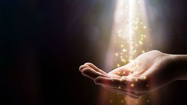 The image depicts an open hand reaching upward, surrounded by a soft, glowing light. From above, beams of light are cascading down, creating a sparkling effect with small particles resembling stars or sparkles. The background is dark, emphasizing the luminous elements in the scene. The overall mood conveys a sense of magic or inspiration.