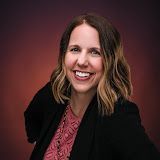 The image shows a person smiling with medium-length, wavy hair that has lighter highlights. They are wearing a black jacket over a pink top with a pattern. The background has a gradient that transitions from dark to light, enhancing the subject's cheerful expression.