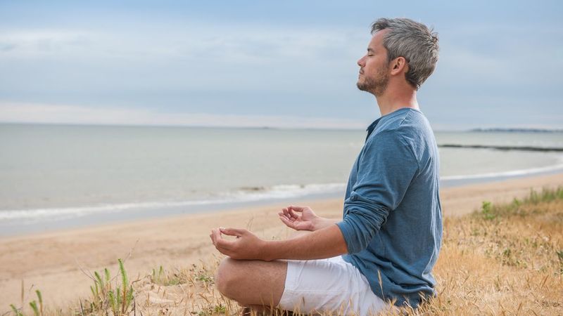 The image shows a man sitting on a sandy beach, meditating. He appears relaxed and is seated in a cross-legged position, facing the ocean. The background features a calm sea and a cloudy sky, creating a serene atmosphere. The man is wearing a blue long-sleeve shirt and white shorts, and is positioned on a patch of grass near the shoreline.