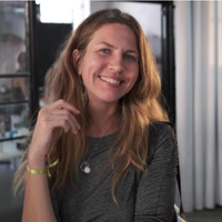 The image depicts a smiling woman with long, wavy hair. She is wearing a dark, fitted top and has a small pendant necklace. Her left hand is raised, and she is holding it near her face. She has a bracelet on her wrist. The background features a bright, airy setting with some light elements and a glass display or partition. The atmosphere appears friendly and casual.