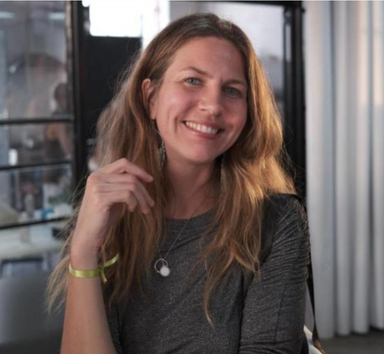 The image depicts a smiling woman with long, wavy hair. She is wearing a dark, fitted top and has a small pendant necklace. Her left hand is raised, and she is holding it near her face. She has a bracelet on her wrist. The background features a bright, airy setting with some light elements and a glass display or partition. The atmosphere appears friendly and casual.